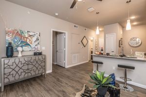 A modern apartment interior featuring a stylish console table with decorative items, a bar-style seating area with stools, and a view of an open kitchen. The walls are painted in a neutral color, and decorative artwork adds color. Soft lighting creates a welcoming ambiance.