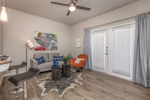 A cozy living room featuring a gray sofa, an orange accent chair, a decorative rug with a zebra pattern, and a wooden coffee table. The space is well-lit with a ceiling fan and has large windows with white blinds. Colorful wall art adds a vibrant touch, and a bar area is visible in the background.