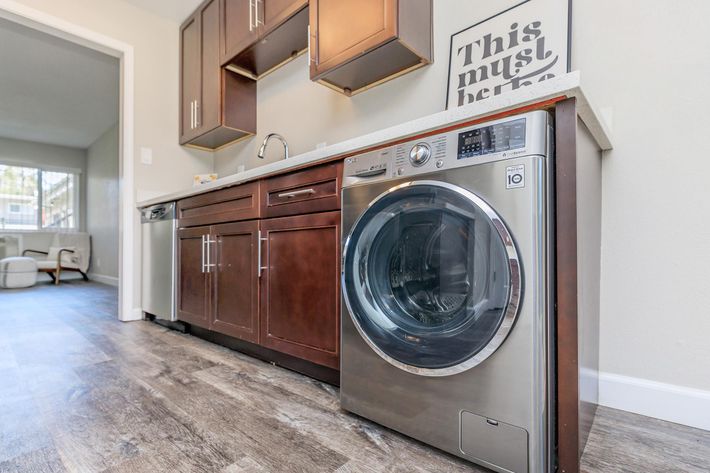 A modern laundry room featuring a silver front-loading washing machine beside dark wooden cabinets. The countertop is light-colored, and there is a framed print on the wall that reads "This must be." The space has a clean and contemporary design with a light-colored floor.