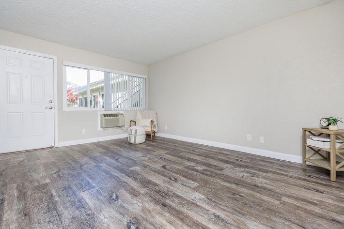 A bright, empty living room featuring light-colored walls, a large window, and wood-like flooring. There's a small chair and a side table against the wall, along with an air conditioning unit. The space is clean and minimalist, creating a welcoming atmosphere.