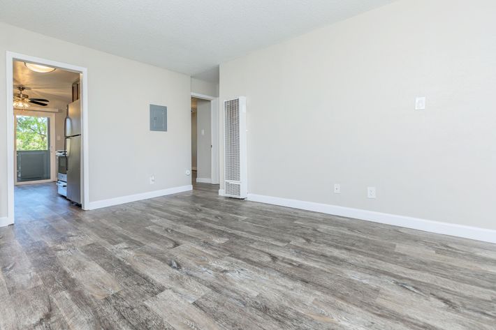 A spacious, light-filled room with a smooth, laminate floor and neutral-colored walls. A doorway on the left leads to a kitchen area, while a vertical heater is visible on the right. Large windows bring in natural light, creating an open and inviting atmosphere.