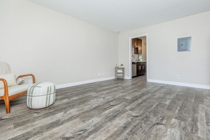 A spacious, empty living room with light-colored walls and modern, laminate flooring. In the foreground, there is a simple wooden chair and a round, patterned pouf. A doorway leads to a kitchen in the background, which features dark cabinetry. Natural light brightens the room, creating an inviting atmosphere.