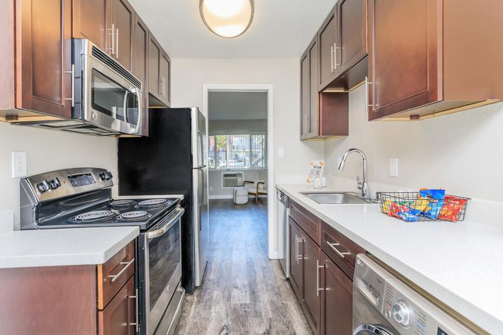 Modern kitchen featuring dark wood cabinetry and stainless steel appliances, including a microwave, oven, and dishwasher. The countertops are light-colored, and there is a sink near a window. A small basket of snacks sits on the counter, and a living area is visible through an open doorway.