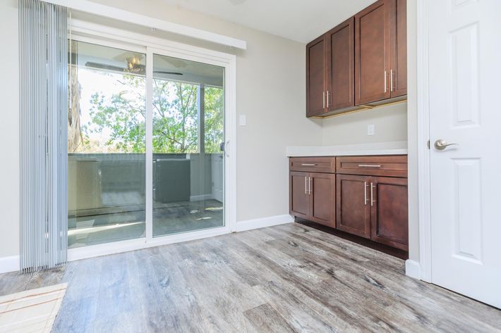 A bright, modern interior view of a room featuring wooden cabinets, a sliding glass door leading to a balcony, and light-colored walls. The flooring is a stylish laminate, and natural light filters in from outside, creating a warm and inviting atmosphere.