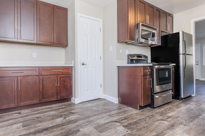 A modern kitchen featuring dark wooden cabinets, stainless steel appliances including an oven, microwave, and refrigerator. The walls are painted light, and the floor has a wood-like finish. There is a closed white door leading to another room, enhancing the space's openness.