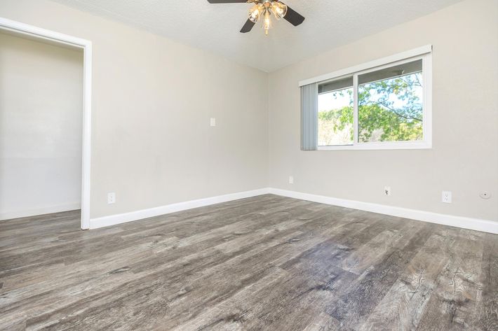 A spacious, empty room featuring light gray walls and a wooden floor. There is a ceiling fan with a light fixture, and a large window allows natural light to enter. One wall has a doorway leading to another area, emphasizing the room's openness and potential for decorating or furnishing.