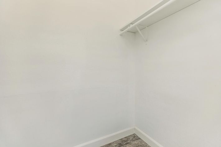 An empty, white-walled closet with a single shelf mounted on the left side. The floor features a wood-like texture, and there are no items stored within the closet, giving it a clean and minimalistic appearance.