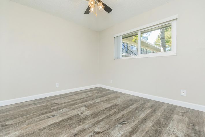 Empty room with light-colored walls and a window allowing natural light in. The ceiling features a black ceiling fan with four light bulbs. The floor is covered in light-colored wood-like laminate. The space appears bright and airy, suitable for various uses.