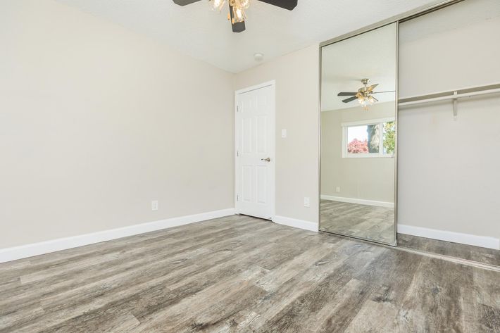 A spacious, empty room featuring light-colored walls, a mirrored closet door, and a ceiling fan. The floor has a wood-like design, and there is a white door to the left leading outside. Natural light fills the space from a window that overlooks greenery outside.