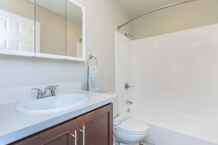 A modern bathroom featuring a white bathtub with a curved shower rod, a sink with a circular basin, and a brown cabinet beneath. The wall is painted light gray, and there is a large mirror above the sink. A towel hangs on a rod on the side, and a white door is visible in the background.