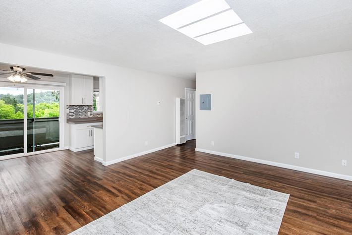 Bright and spacious interior of a modern living area with wooden floors and a large window allowing natural light. The room features a minimalist design with neutral walls and a light-colored rug on the floor. An open doorway leads to a kitchen area in the background with sliding doors and greenery visible outside.