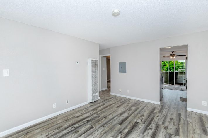 A spacious, well-lit living area featuring light gray walls and laminate flooring. There is a wall-mounted air conditioning unit and a small electrical panel visible. The room opens to a bright area with a ceiling fan and large windows, suggesting a connection to an outdoor balcony or patio.