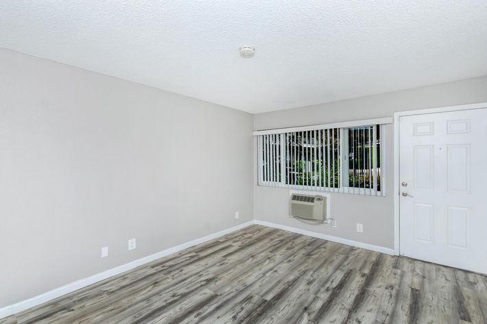A small, empty room with light gray walls and modern laminate flooring. There is a window with vertical blinds allowing natural light, and an air conditioning unit mounted below. A white door is visible on the right side of the room, creating a bright and airy atmosphere.