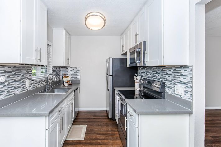 A modern kitchen featuring dark wooden cabinets, a stainless steel refrigerator, and an electric stove. The countertop is light-colored, complementing the sleek design. The kitchen leads to a bright area visible in the background, with large windows allowing natural light.