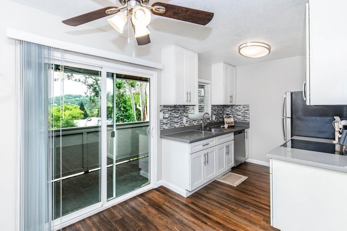 A modern kitchen featuring dark wood cabinets, a stainless steel sink, and a washing machine. The walls are painted white, and a ceiling fan is visible. The flooring is light-colored laminate. An open doorway leads to another room, and natural light enters through a window.