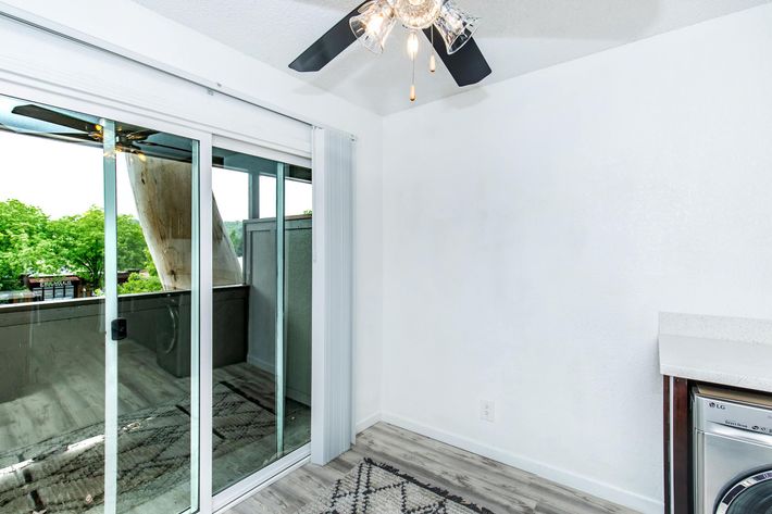 A bright, modern room featuring a stylish ceiling fan, large glass sliding door with balcony access, and light-colored walls. The floor is sleek and wooden, with a rug placed near a washing machine. The space is well-lit with natural light coming through the sliding door, and greenery is visible outside.