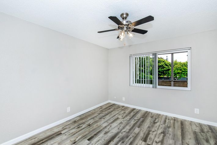 A bright, empty room featuring light-colored walls and wood-like flooring. It has a ceiling fan with a light fixture, and a window with vertical blinds allows natural light in. Outside the window, greenery is visible. The space is clean and ready for furniture arrangement.