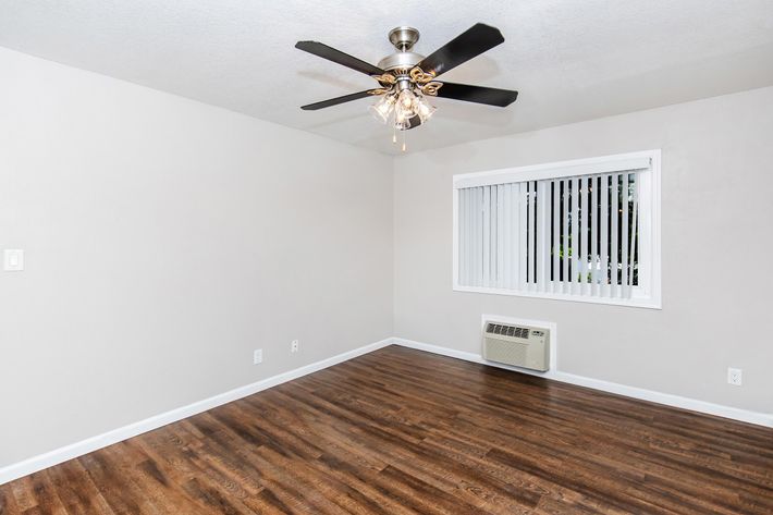 An empty room featuring light gray walls and a ceiling fan with a light fixture. The floor is covered in laminate wood planks. A doorway on the left leads to another space that is partially visible. The overall ambiance is bright and clean, suitable for various uses.
