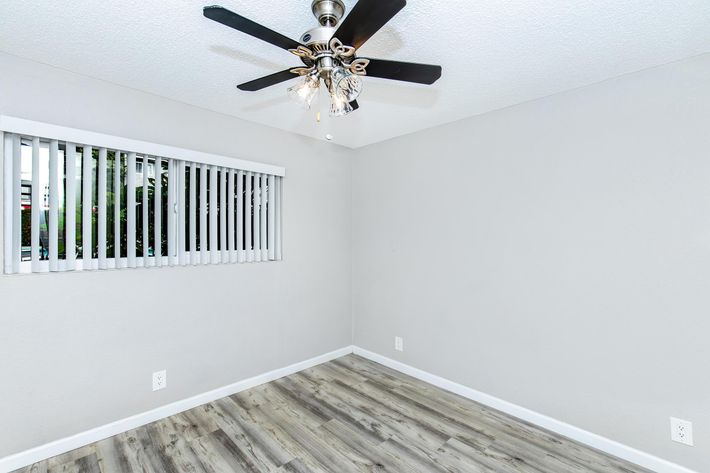 An empty room with light gray walls and a wooden floor. A ceiling fan with light fixtures hangs from the ceiling. A window with horizontal blinds provides natural light. The space appears ready for furnishing and personalization.