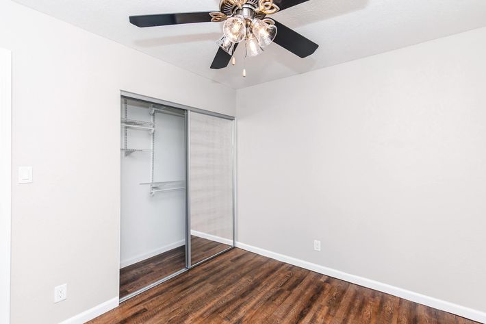 A clean, modern bedroom featuring a ceiling fan with light fixture, light gray walls, and dark wood-like laminate flooring. On one side, there's a mirrored sliding closet door, revealing a simple storage solution with shelves. The space is well-lit and uncluttered, creating a serene environment.