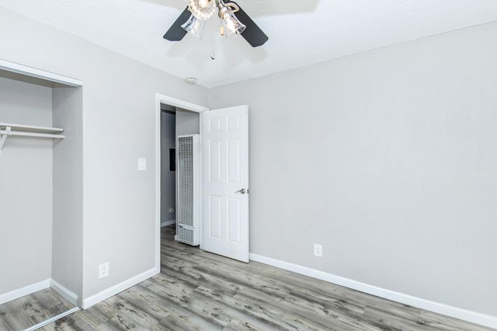 A light grey room featuring a ceiling fan with a chandelier style light. There’s a white door leading to another room, a closet space on the left with no doors, and light wood-like flooring. The walls are bare, creating a clean and simple aesthetic.