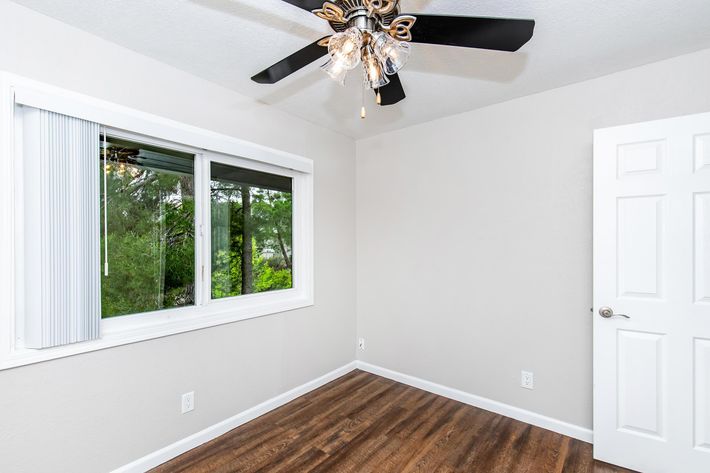 A minimalist room with light gray walls and wood-like flooring. A ceiling fan is visible, and there's a large mirrored closet with sliding doors along one wall. The space is well-lit and appears clean and empty, suitable for a bedroom or office.