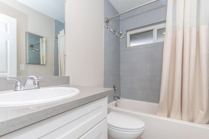 A clean, modern bathroom featuring a white tub with a shower, gray tiled walls, a simple vanity with a round sink, a large mirror, and a light-colored countertop. The walls are painted white, and there's a shower curtain in a neutral shade.