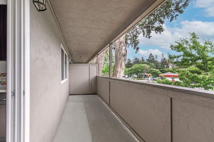 A wide balcony featuring a concrete floor, partially enclosed by a beige wall. A railing runs along one side, offering a view of trees and buildings outside. Natural light filters in through large windows, and the sky is clear with a few clouds visible.