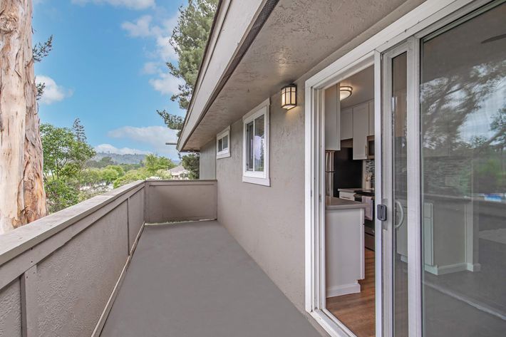 View of a balcony with a gray floor and sliding glass door, overlooking greenery and rolling hills. The exterior features light-colored walls and a few trees in the background under a partly cloudy sky. The balcony is spacious, providing a serene outdoor space adjacent to a modern interior.