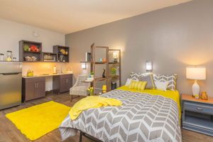 A cozy studio apartment featuring a bed with a geometric gray and yellow comforter, a bedside lamp, and a small seating area. The kitchenette includes a fridge and modern cabinetry, while a bright yellow area rug adds a cheerful touch to the space. Walls are painted in a neutral tone, creating a warm atmosphere.