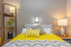 A cozy bedroom featuring a yellow and gray patterned bedspread with decorative pillows. There are two lamps, one on a bedside table and another on a bookshelf, along with a shelf displaying a vase of flowers and some books. The walls are painted a soft gray, creating a warm and inviting atmosphere.