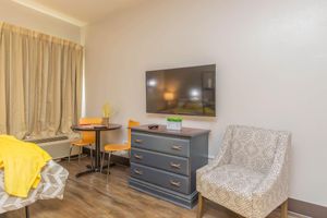 A cozy hotel room featuring a gray dresser with drawer handles, a mounted flat-screen TV on the wall, a small round dining table with two orange chairs, and a patterned armchair. A bed with a yellow blanket is visible, along with large curtains letting in natural light.