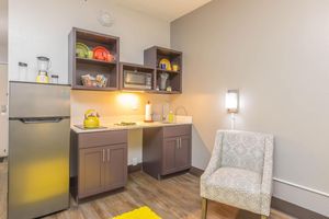 A modern kitchenette featuring a stainless steel refrigerator, microwave, and dark cabinets. The countertop displays colorful dishes and a kettle. A small, decorative chair sits nearby on a yellow rug, with a wall-mounted light providing soft illumination. The overall aesthetic is clean and inviting.