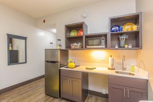 A small kitchen area featuring a stainless steel refrigerator, a microwave, and a sink. Open shelves display colorful plates and kitchen utensils. There is a mirror on the wall, and the flooring is a light wood. The space is well-lit and designed in neutral tones.