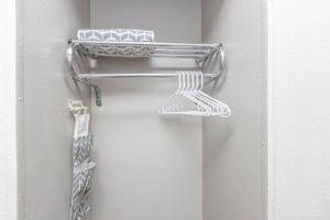 A small closet with a light gray wall. It features a metal shelf with a folded gray and white towel on top, underneath which hangs several white plastic hangers. A wall-mounted organizer with gray patterns holds additional storage items, enhancing the closet's functionality.
