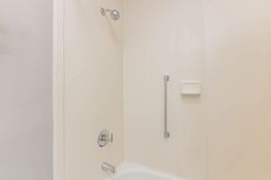 A clean, simple shower stall featuring a white tiled wall, a chrome showerhead, a grab bar for safety, and a soap dish. The bathtub is visible at the bottom, with a smooth, white finish. The overall appearance is bright and well-maintained, with neutral colors creating a spacious feel.