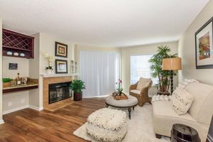 Cozy living room featuring a beige sofa and a patterned armchair. A round coffee table with decorative items sits on a plush rug. A fireplace is complemented by a plant and a floor lamp. Large windows provide natural light, and elegant wall art adds charm to the space.