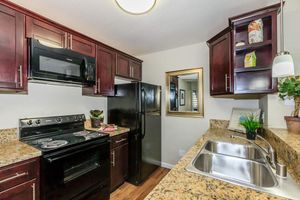 A modern kitchen featuring dark wood cabinets, stainless steel appliances including a microwave and black refrigerator, and a granite countertop. The double sink is positioned under a light fixture, and there's a framed mirror on the wall. Decorative plants and a few items are on the countertop.