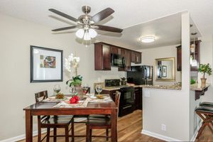 A modern kitchen and dining area featuring a wooden dining table set for two, with decorative plates and glasses. The kitchen has dark cabinets, stainless steel appliances, and a bar counter with stools. A ceiling fan provides airflow, and potted plants add a touch of greenery. Walls are light-colored, creating a warm atmosphere.