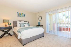 A cozy bedroom featuring a king-sized bed with decorative pillows, a stylish nightstand with a lamp, and a chic dresser. The walls are painted in soft neutral tones, and large sliding glass doors lead to a balcony with palm trees visible outside, enhancing the inviting ambiance of the space.