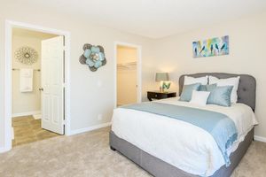 A bright and cozy bedroom featuring a queen-sized bed with light blue and white bedding. There is a bedside table with a lamp, and decorative wall art above the bed. A door leads to a bathroom, and another door opens to a closet. The room has light-colored walls and soft carpet, creating an inviting atmosphere.