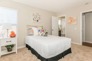 A cozy bedroom featuring a neatly made single bed with colorful patterned pillows. A small side table with a lamp is next to the bed, and there are two decorative fish paintings on the wall. The room has light beige walls and carpeted flooring, with a partially visible bathroom door in the background.