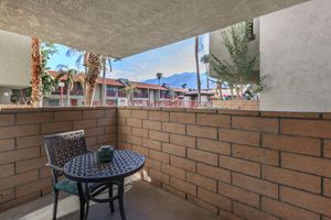 A cozy balcony setting featuring a round table with a decorative centerpiece and a single chair. The view includes palm trees and a distant mountain range, with buildings visible in the background under a clear sky. The walls are made of light-colored bricks, creating a warm and inviting atmosphere.