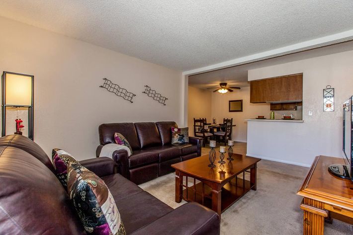 A cozy living room featuring a dark leather sofa with decorative cushions, a wooden coffee table with candles, and a flat-screen TV. In the background, there is a dining area with a ceiling fan and a kitchen bar visible. The walls are adorned with minimalist decor, creating a warm and inviting atmosphere.