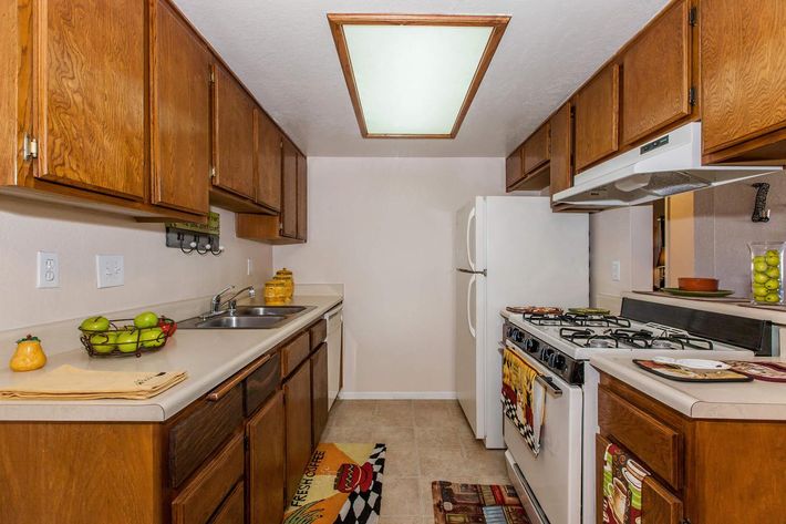 A modern kitchen featuring wooden cabinetry, a double sink, and a white refrigerator. The room includes a gas stove with an oven, colorful kitchen mats, and decorative fruit in a bowl on the counter. A ceiling light fixture provides illumination in the space.