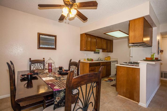 A cozy kitchen and dining area featuring a brown wooden table set with dishes, a ceiling fan, and a well-equipped kitchen with wooden cabinets. The space includes a small dining table, countertop with appliances, and a warm, inviting ambiance. Bright lighting enhances the room's cheerful atmosphere.