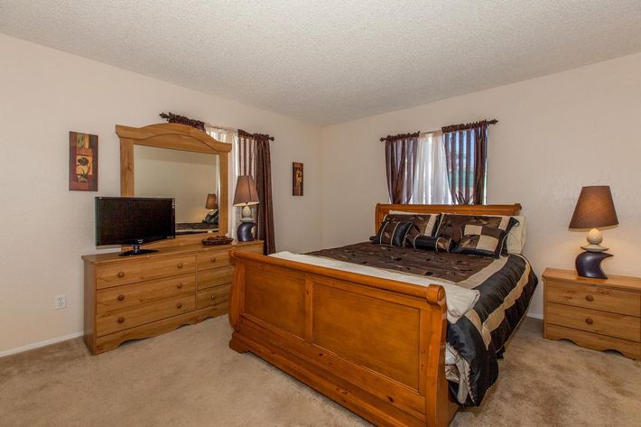 A cozy bedroom featuring a wooden bed with a dark patterned comforter, flanked by matching nightstands and a wooden dresser. A television sits on the dresser, and a large mirror reflects the room. The walls are light-colored, and there are two windows with sheer curtains letting in natural light.