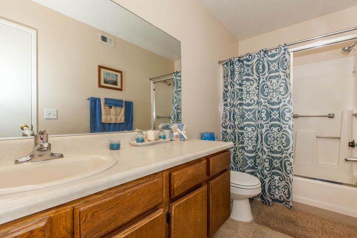 A clean and organized bathroom featuring a beige wall, wooden cabinets, and a white sink. A large mirror reflects the space, while a blue decorative shower curtain adds color. The countertop is adorned with various toiletries, and there's a towel rack with a folded blue towel. A toilet is visible, alongside a shower stall.