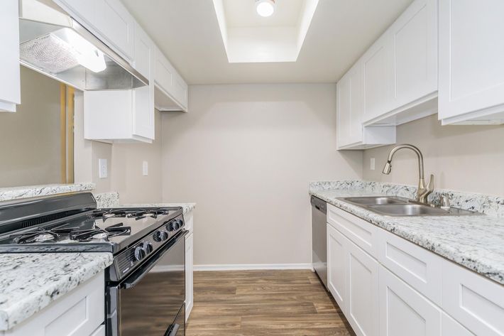 A modern kitchen featuring white cabinetry, granite countertops, a stainless steel sink, and a gas stove. The room is well-lit with a skylight overhead and has a warm wood-style floor, creating a clean and inviting atmosphere.