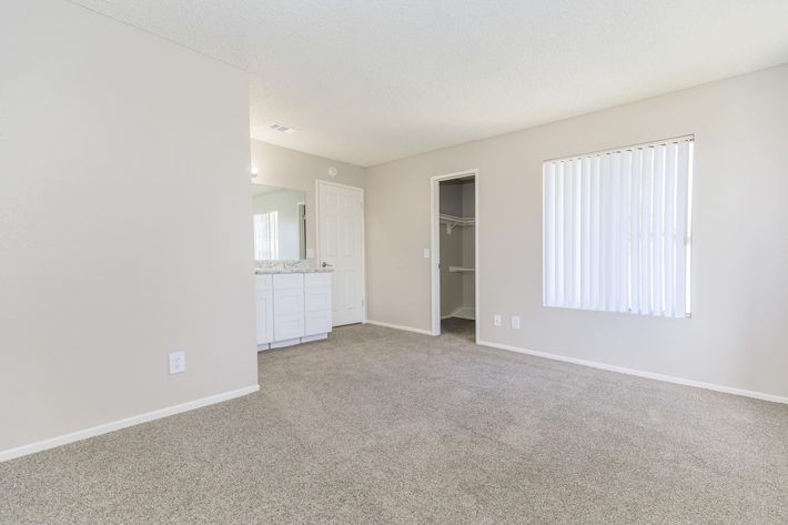 A spacious, light-colored room with beige carpet and neutral walls. It features a bathroom area with a double sink vanity, a closet to the right, and a window with vertical blinds allowing natural light. The overall atmosphere is clean and minimalistic.